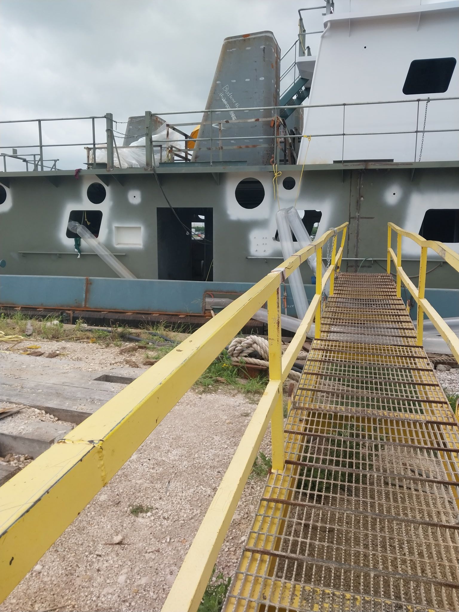 Yellow metal walkway leading to a gray ship’s deck with railings and hatches at a dock