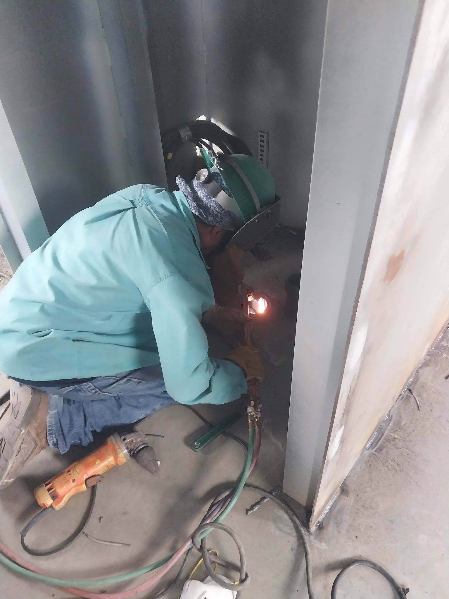 Worker in teal jacket welding inside a metal enclosure, wearing a helmet and crouching near sparks.