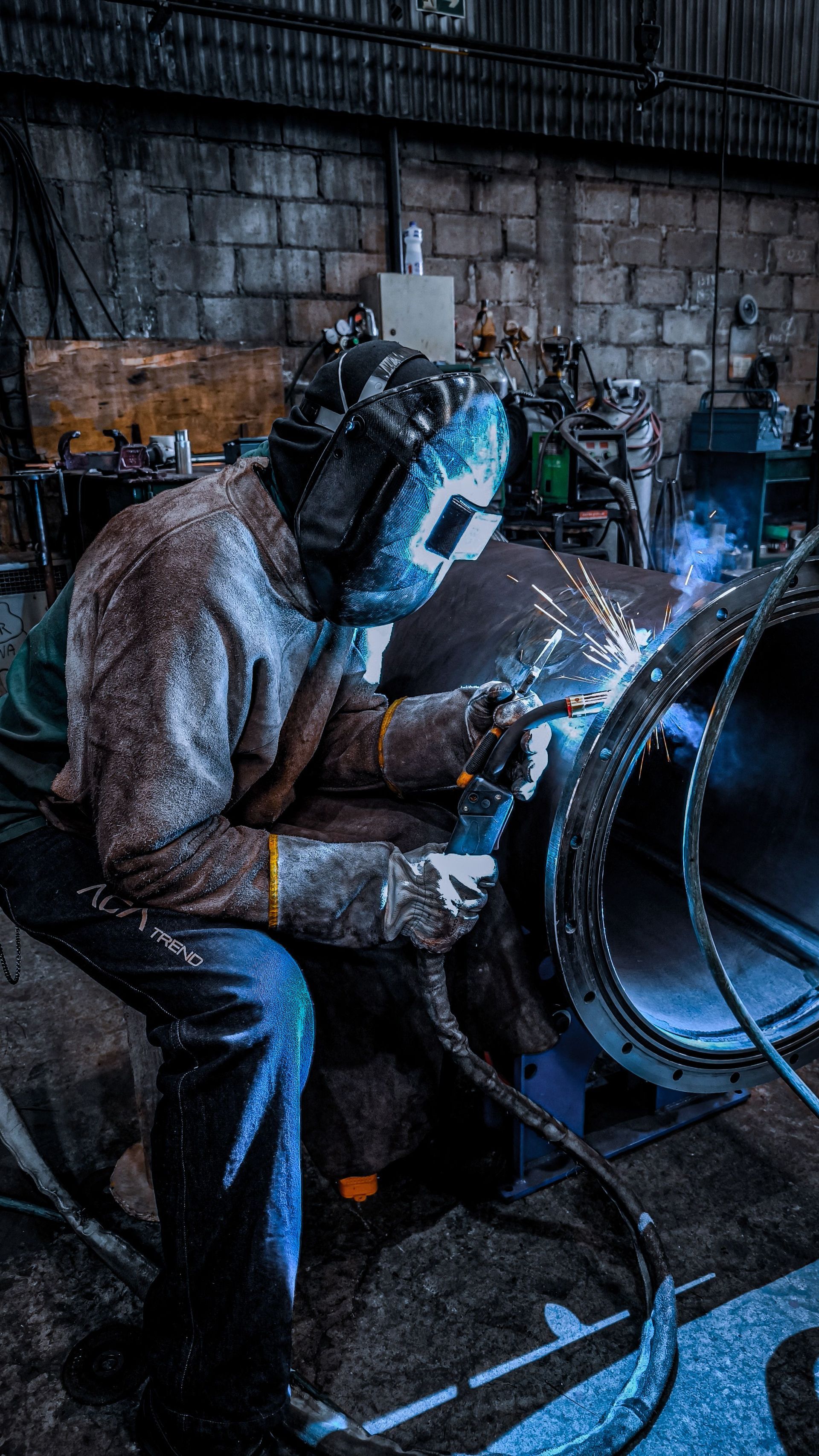 Worker welding a large metal pipe in a dim industrial workshop, sparks flying.