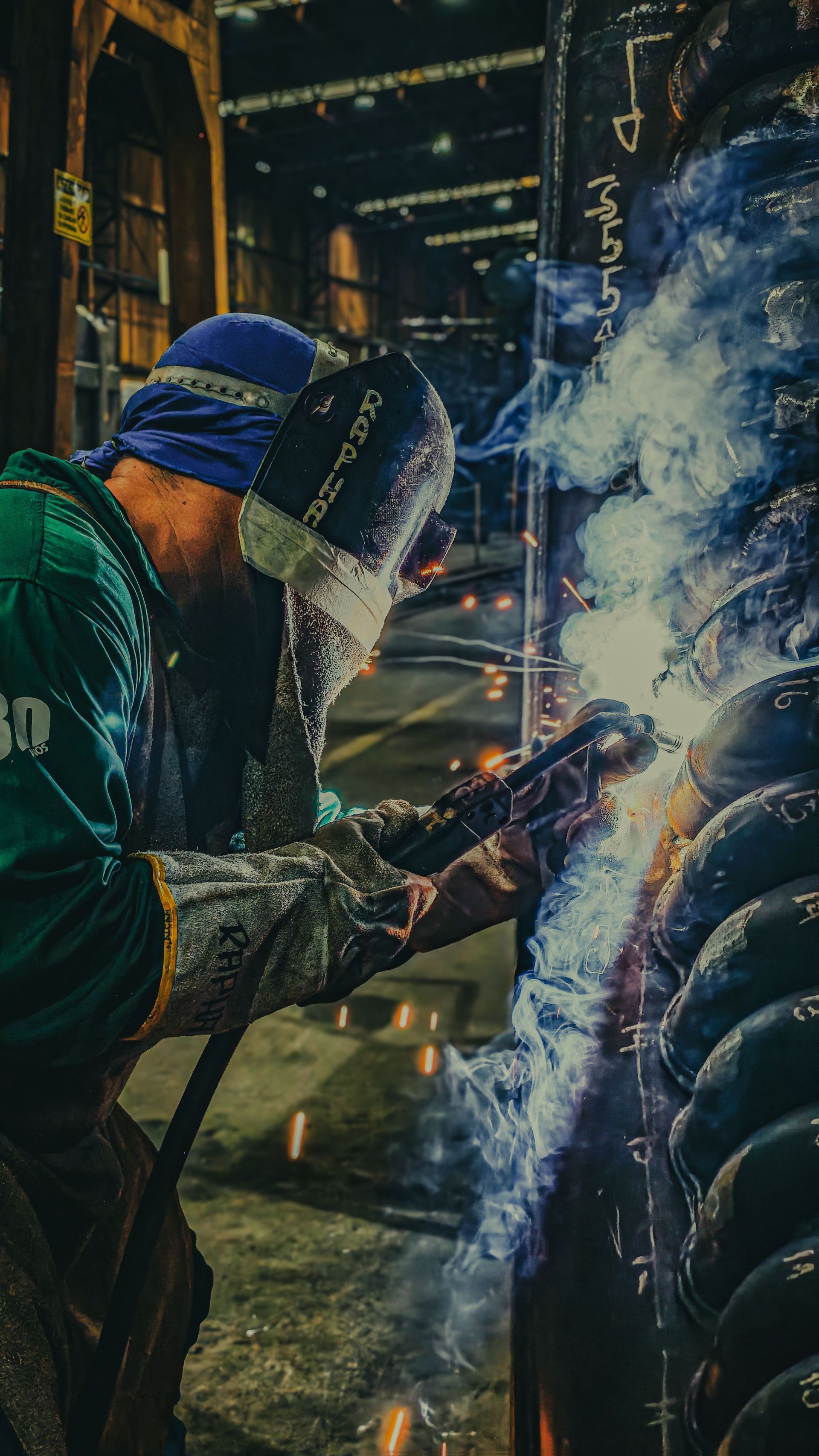 Welders in protective gear working on a large metal structure, sparks and blue smoke flying.