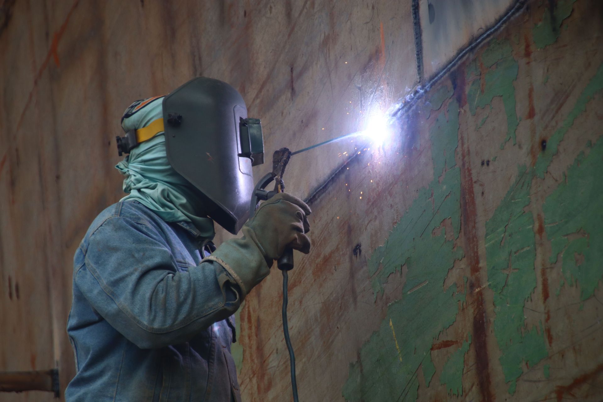 Worker in protective gear welding on a large metal wall, with a bright spark in a narrow industrial space.
