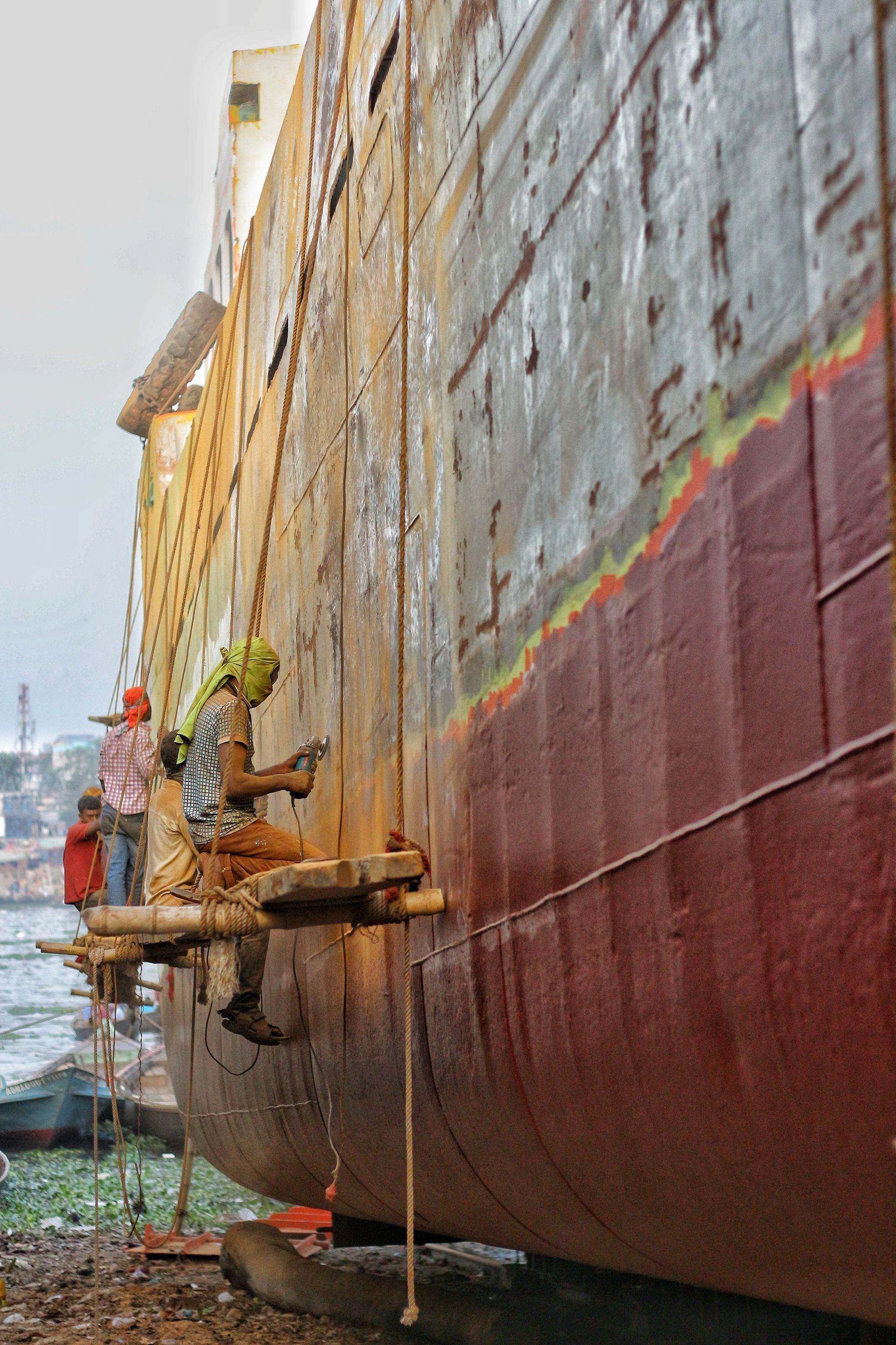 Workers on a ship hull using a suspended platform for painting and maintenance.