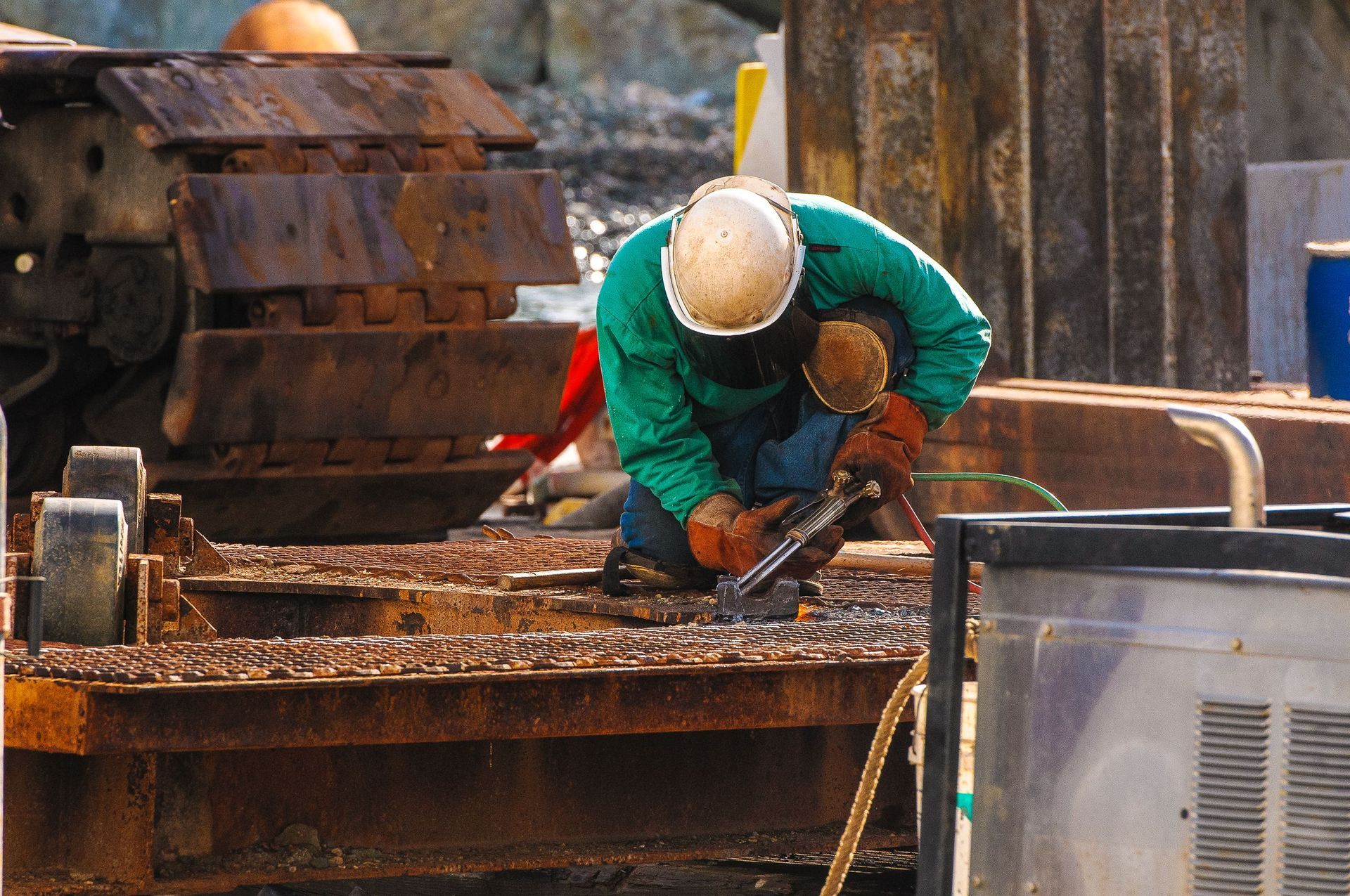 Worker in green jacket and white hard hat bends over a rusty industrial machine outdoors.