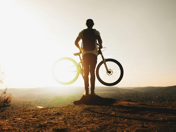Cyclist silhouetted at sunset, standing with bike on a hilltop overlook.