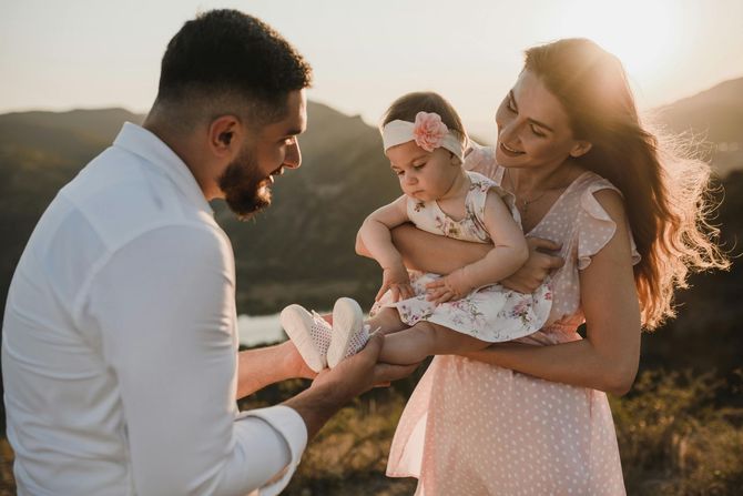 Family outdoors at sunset, parents smiling at a baby in a pink dress in a sunlit field