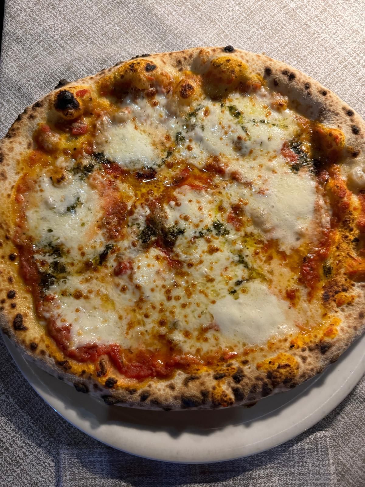 Pizza al formaggio con crosta dorata su un piatto bianco appoggiato su una tovaglia grigia.