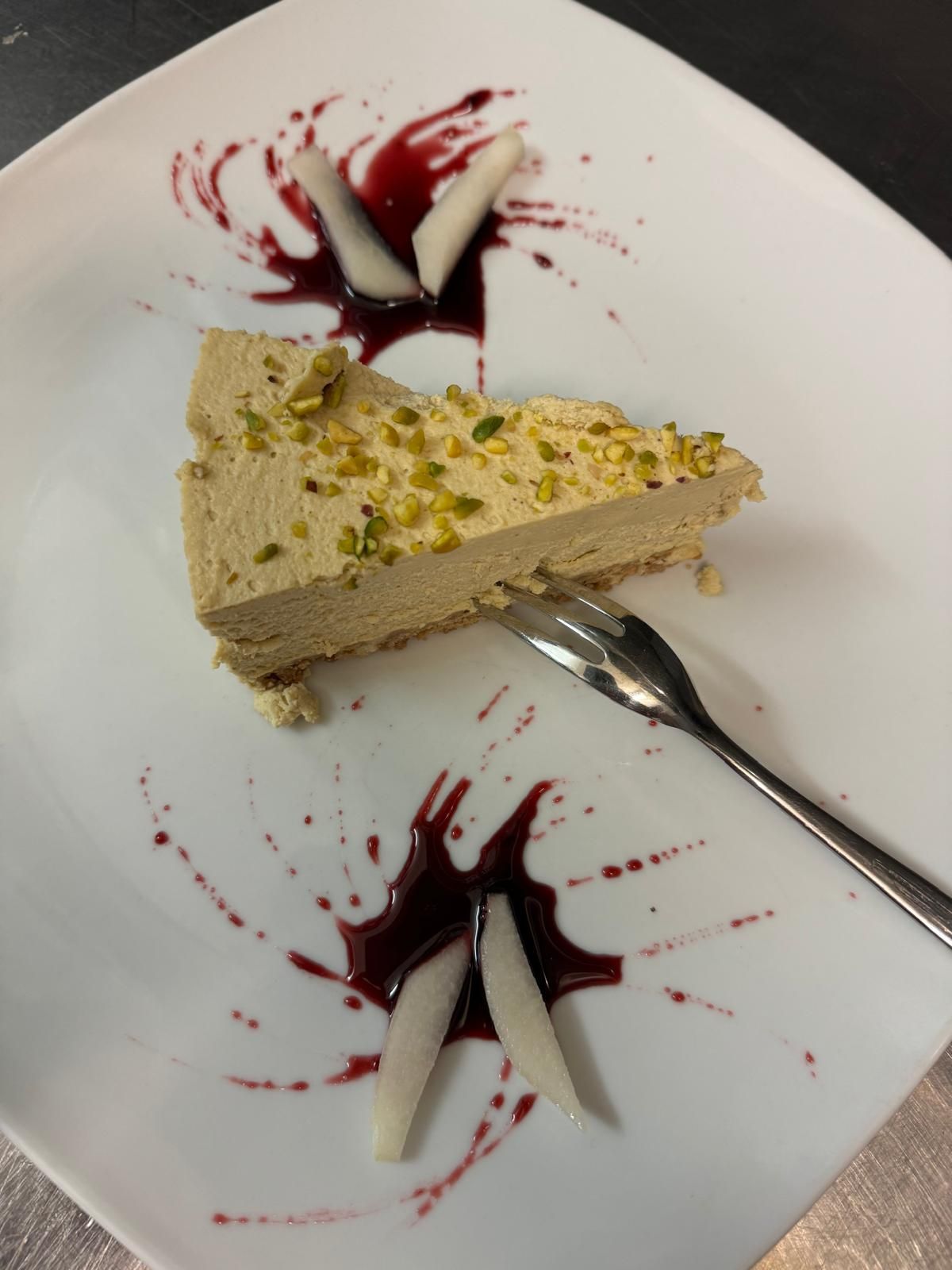 Fetta di torta al pistacchio su un piatto bianco con salsa ai frutti di bosco e ciuffi di panna.