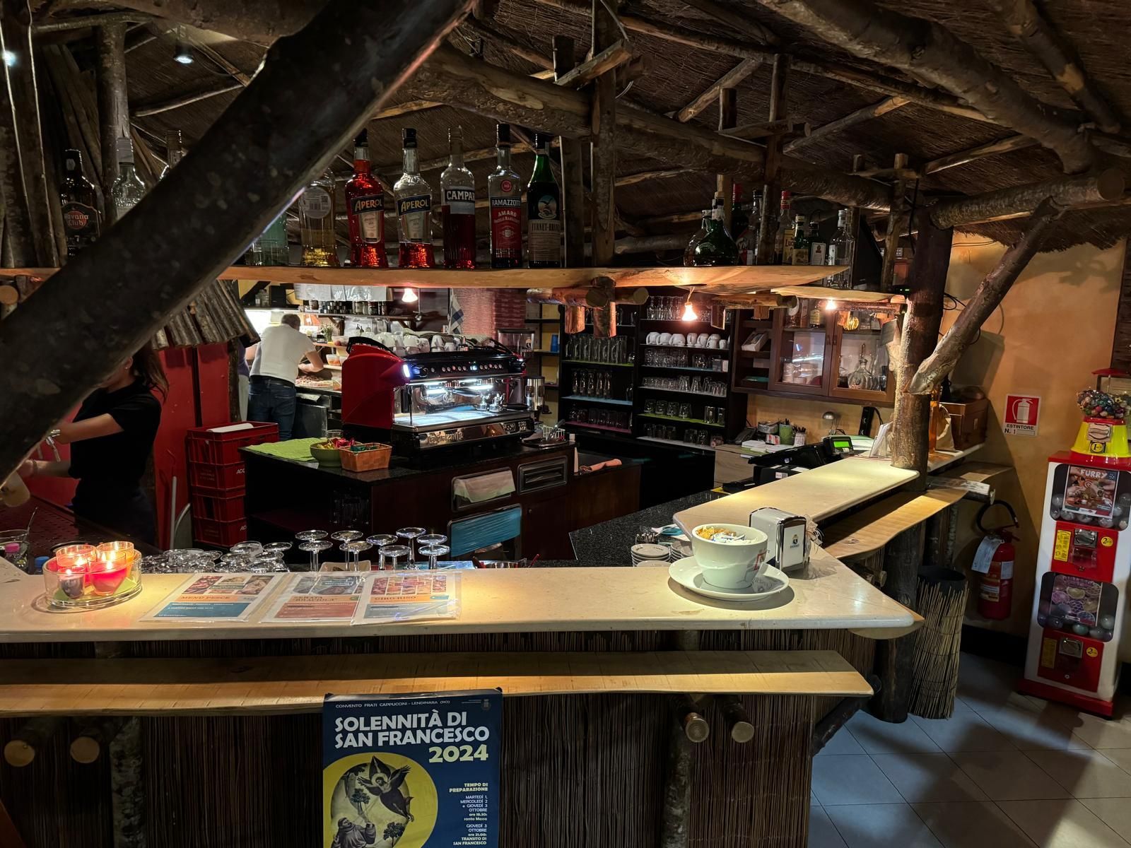 Interno rustico di un bar con un lungo bancone, scaffali pieni di bottiglie e travi di legno a vista sotto un tetto di paglia.
