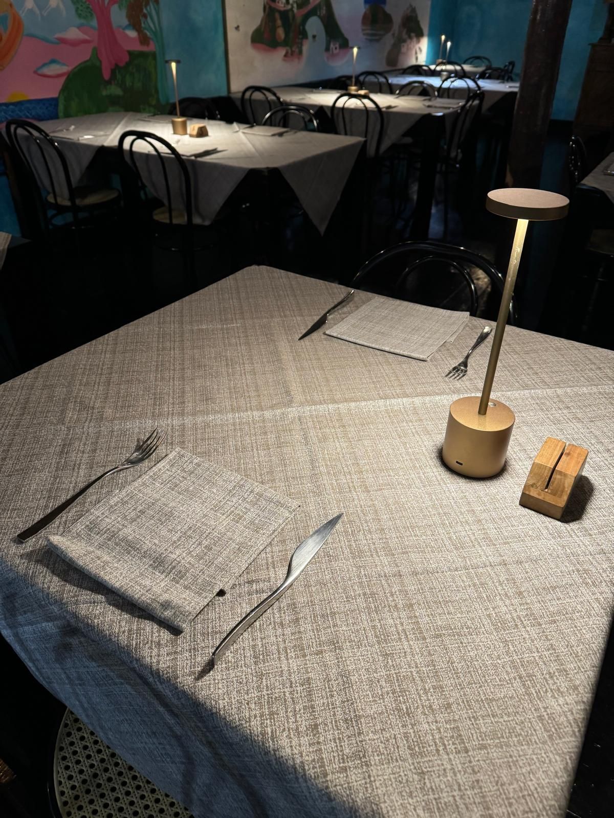 Tavolo di ristorante in penombra con una tovaglia grigia, tovaglioli piegati, una lampada e tavoli sullo sfondo apparecchiati per la cena.