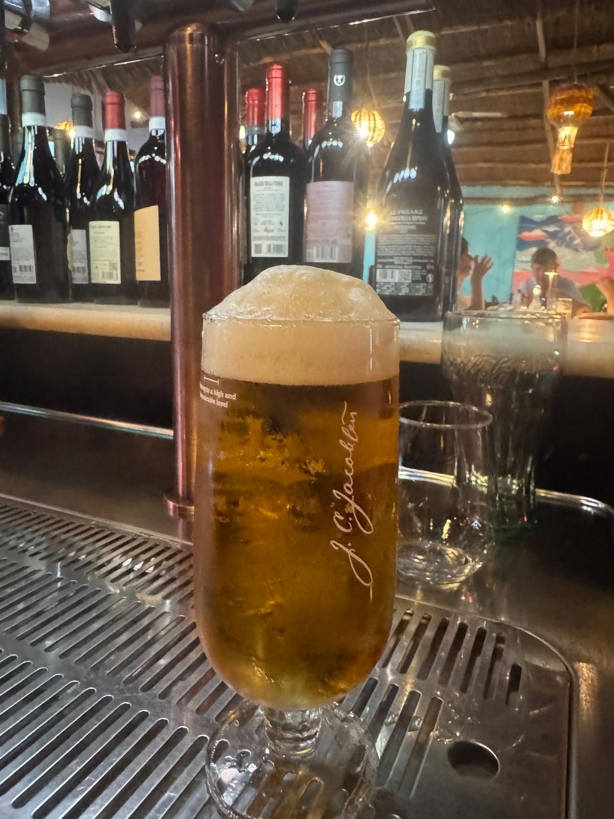 Una pinta di birra con la schiuma addosso, appoggiata sul bancone del bar, con bottiglie di vino allineate dietro.