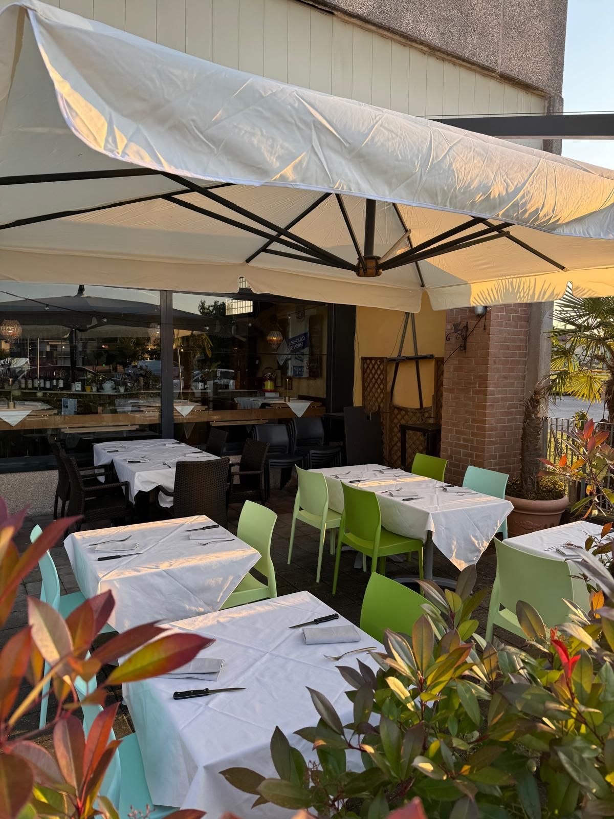 Dehor esterno del caffè con tavolini con tovaglie bianche, sedie verdi e un grande ombrellone, immerso nella calda luce del sole.