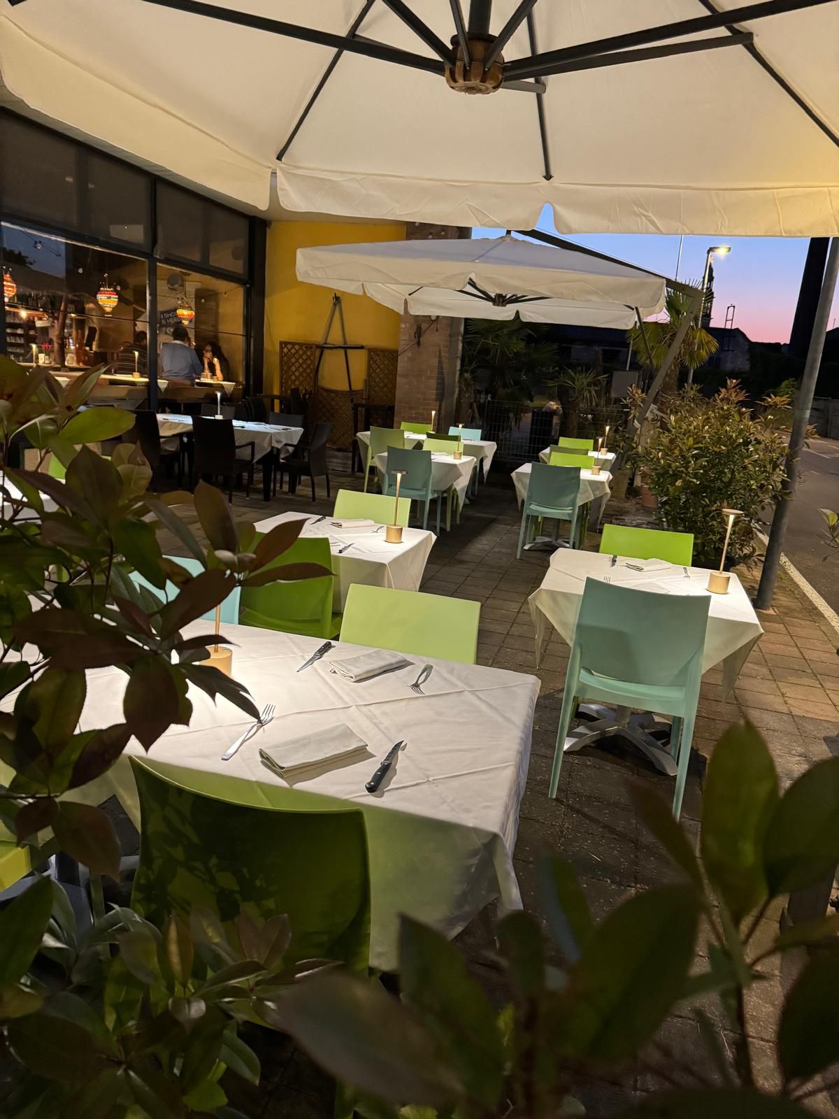 Dehor esterno del caffè con tavolini bianchi, sedie verdi e grandi ombrelloni al tramonto.