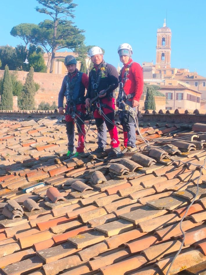 Tre alpinisti con il casco in piedi su un tetto in terracotta, con il campanile di una chiesa sullo sfondo