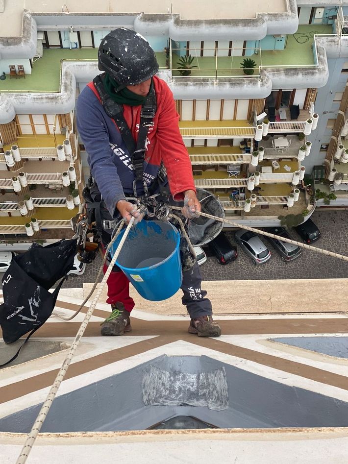 Un operaio con abiti rossi e blu navy versa acqua da un secchio blu su una struttura sul tetto.