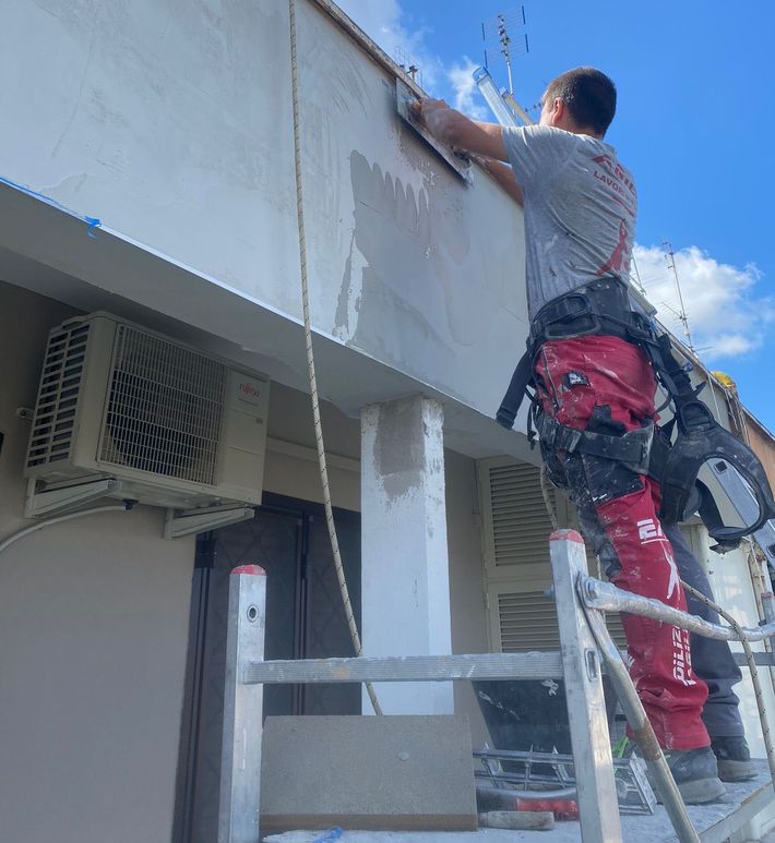 Un operaio su una scala ripara la parete esterna di un edificio sotto un cielo azzurro.