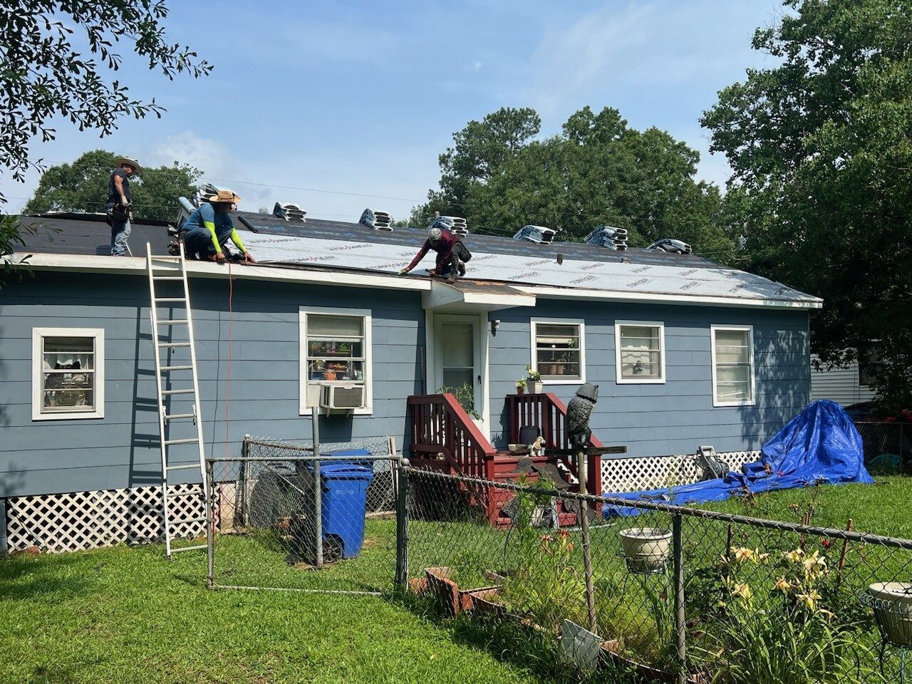Workers repairing a gray house roof, with ladders, tools, and a fenced yard in front