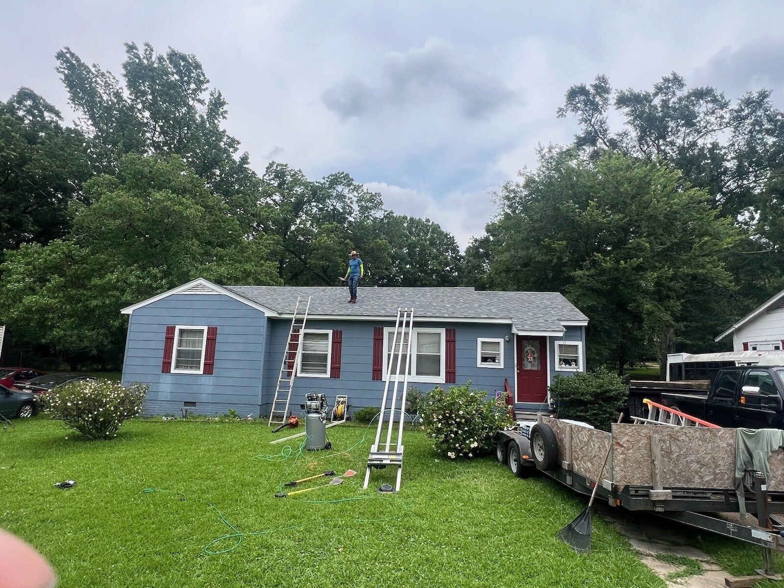 Blue single-story house with white trim and red shutters, a green lawn, and a trailer in the driveway.