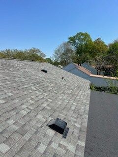 Gray shingle roof with two vents and blue sky above trees and neighboring roofs