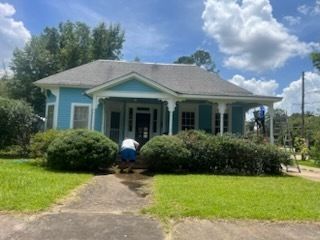 Blue house with porch, person washing sidewalk, green lawn and bushes. Cloudy sky.