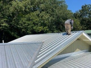 Worker on a silver metal roof against trees and blue sky.