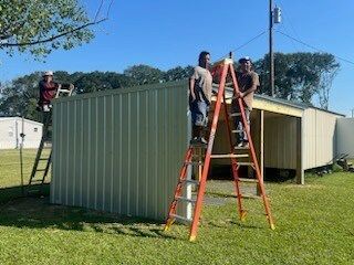 Three people on ladders working on the roof of a green metal building outdoors.