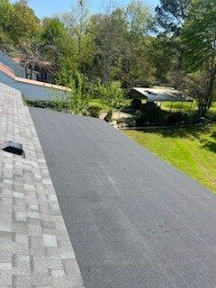 Residential rooftop with dark shingles, skylights, and surrounding trees on a sunny day