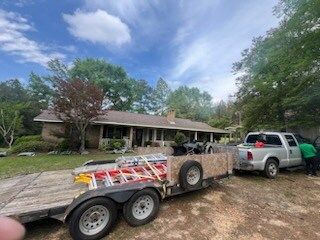 Two pickup trucks and a trailer parked in front of a single-story house on a dirt driveway.