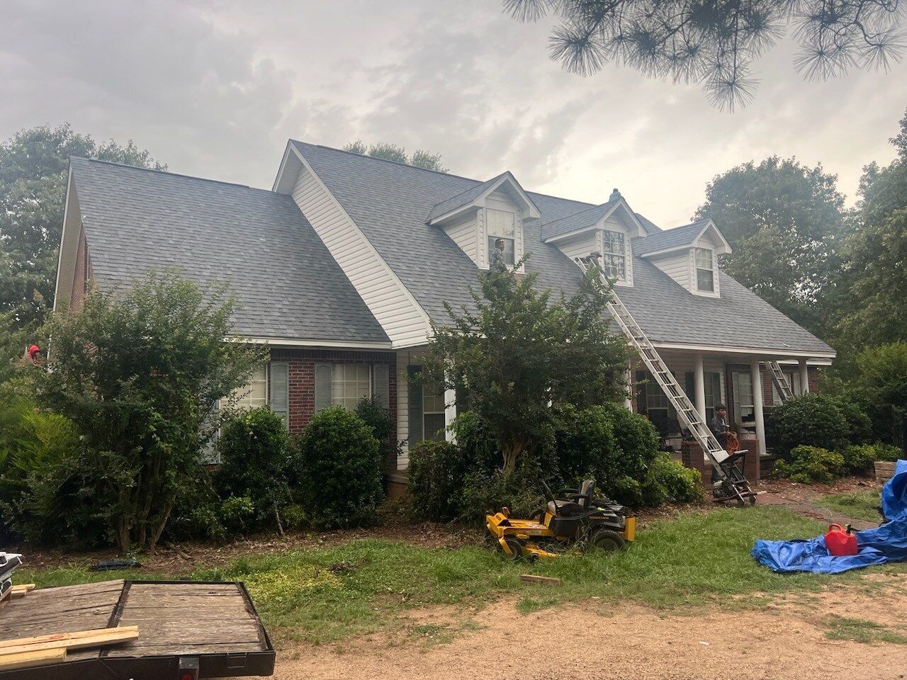 Damaged gray-roofed house with overgrown yard, fallen branches, and scattered debris after a storm