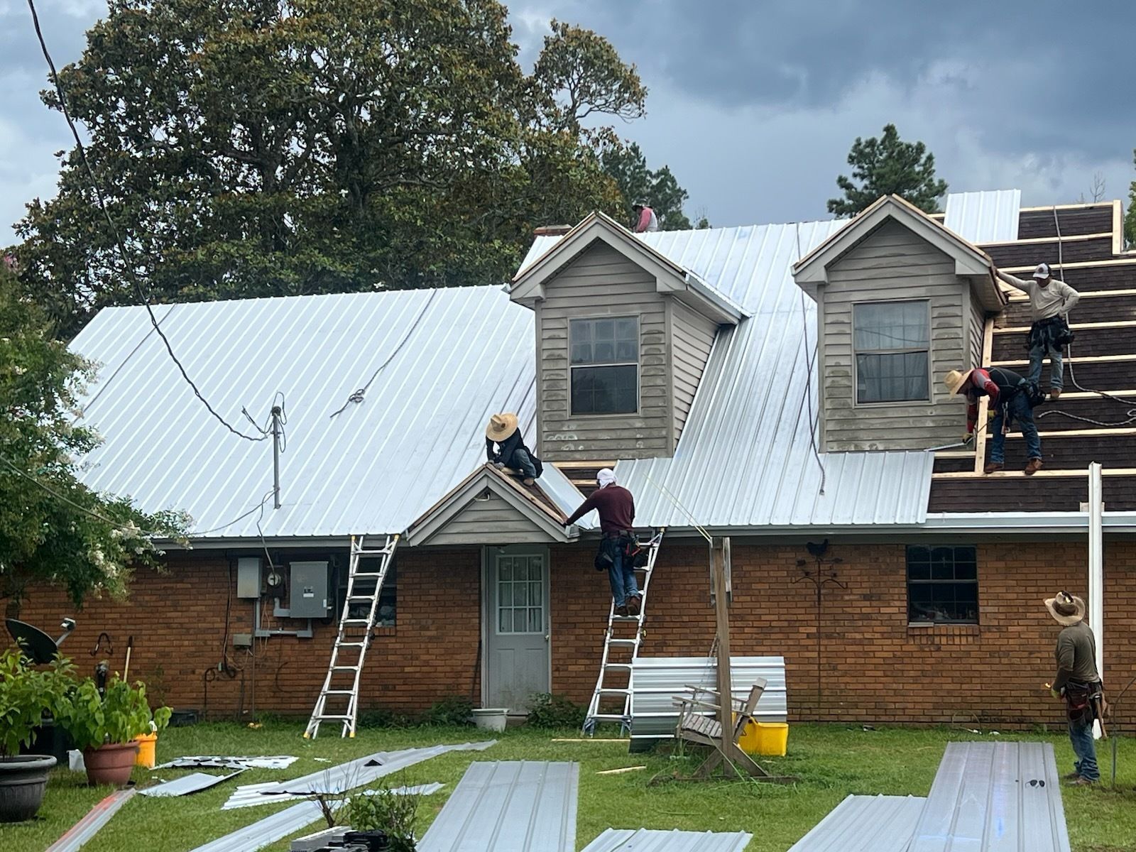 Workers repairing a white metal roof on a brick house, with ladders and scaffolding in a yard.