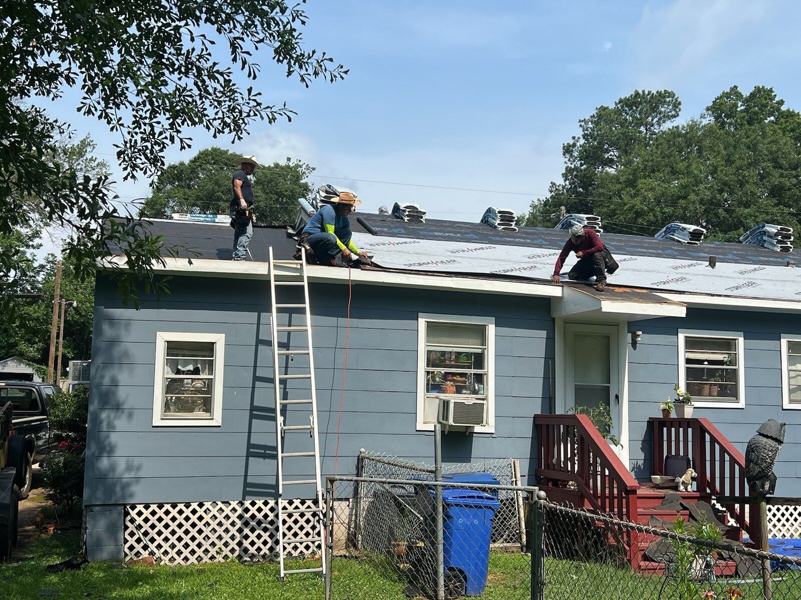 Workers on a blue house roof after storm damage, with a ladder and debris nearby.