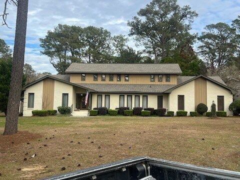 Large beige house with dark trim, front lawn, and trees under a partly cloudy sky.