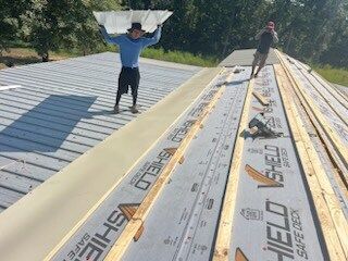 Two workers carrying roofing panels on a sloped roof, with materials spread along the ridge.