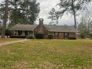 Single-story brick house with a chimney, front porch, and large yard surrounded by tall trees
