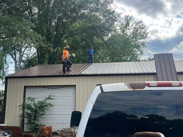 Two workers on a metal roof repairing a beige garage under cloudy trees