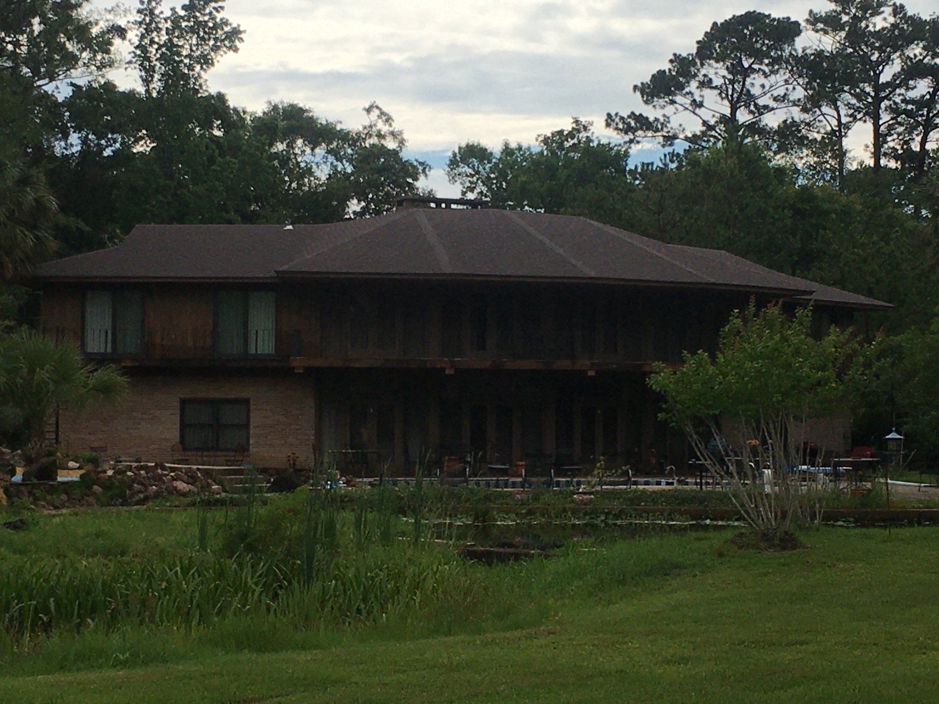 Large house with a dark roof beside a grassy yard and trees under a cloudy sky