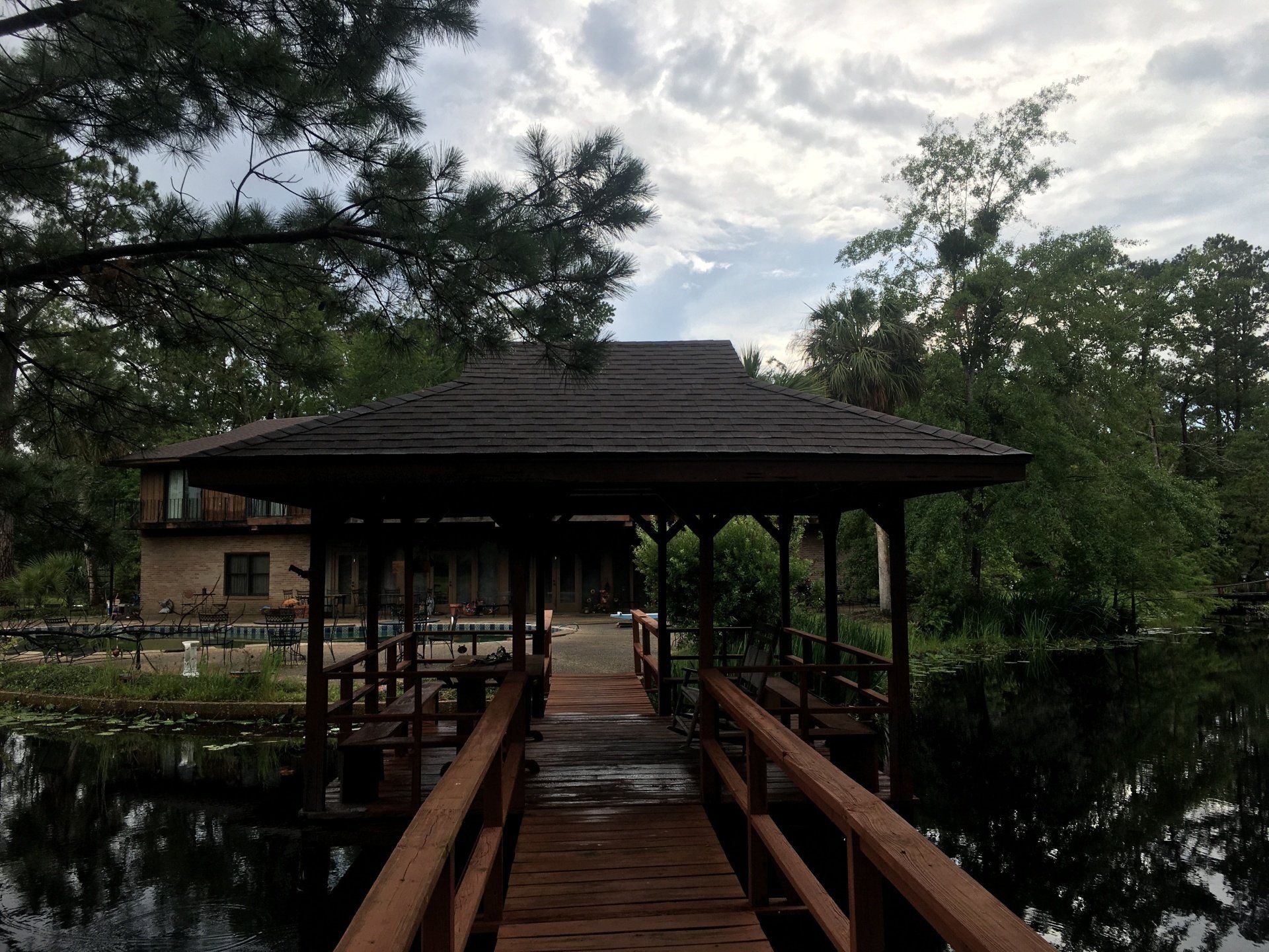 Wooden bridge leading to a covered pavilion over still water, surrounded by trees under a cloudy sky