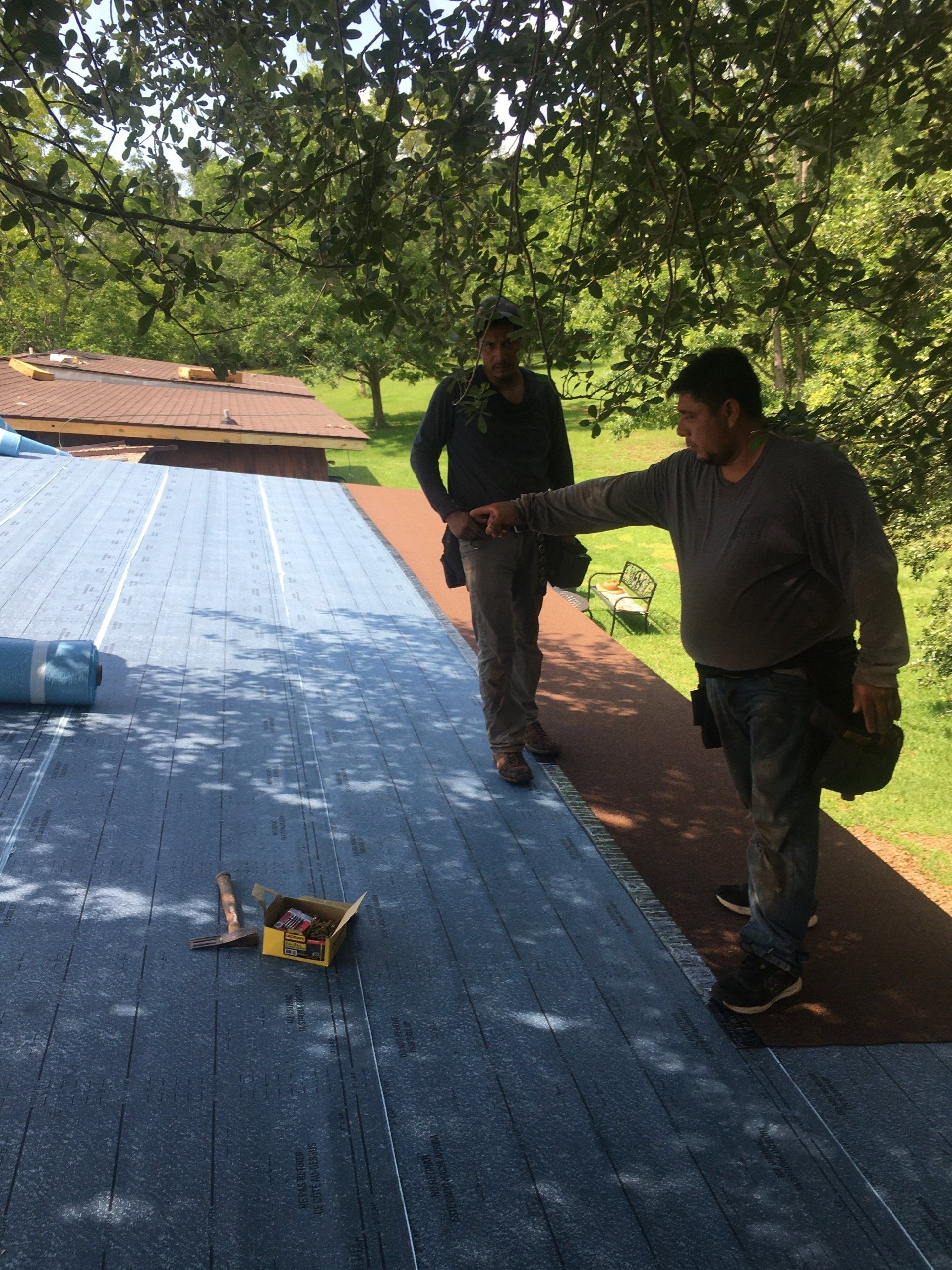 Two workers stand on a blue tarp-covered roof near tools and trees in the background