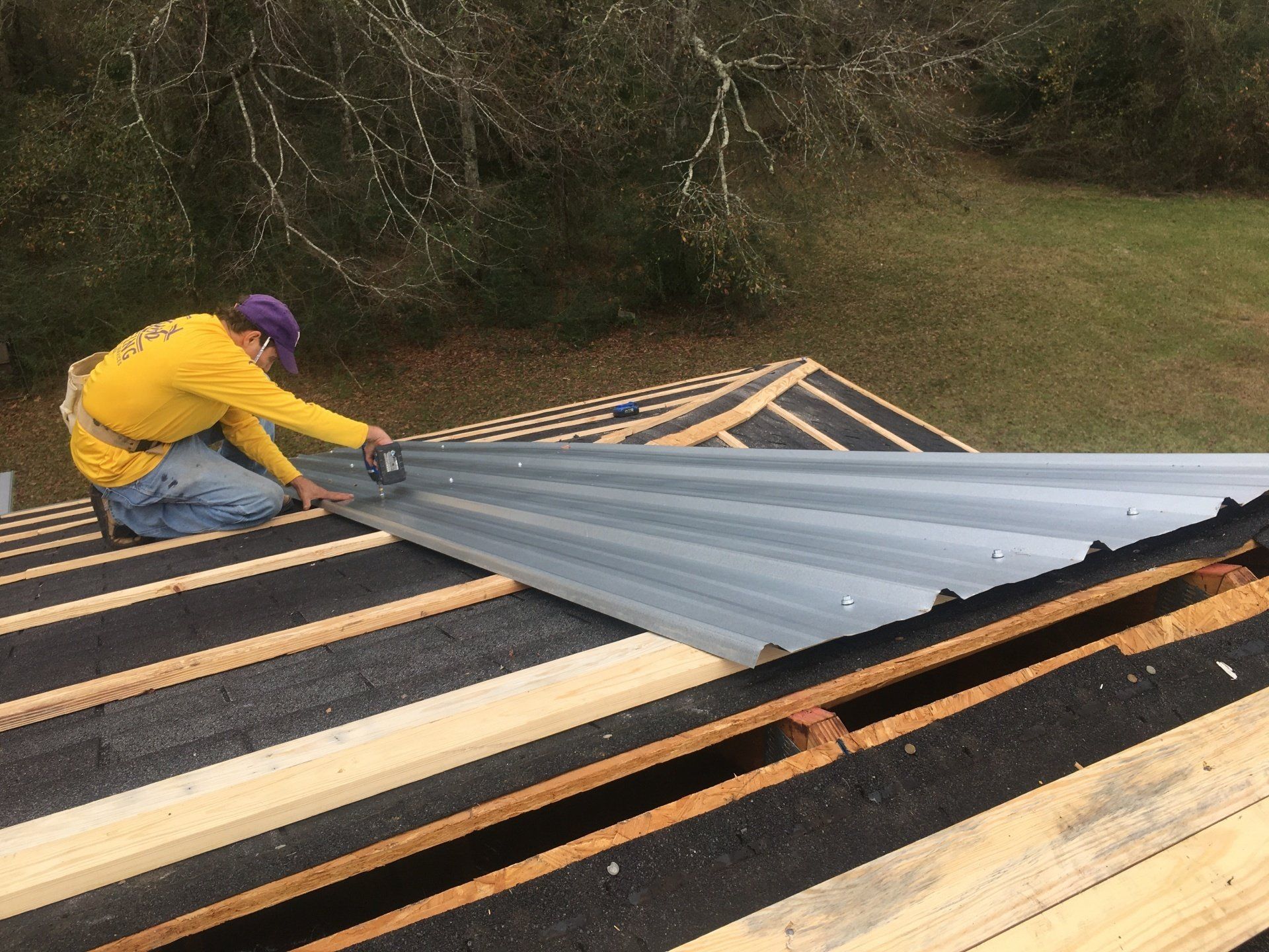 Worker in yellow jacket and purple hat installing corrugated metal roofing on a roof frame outdoors