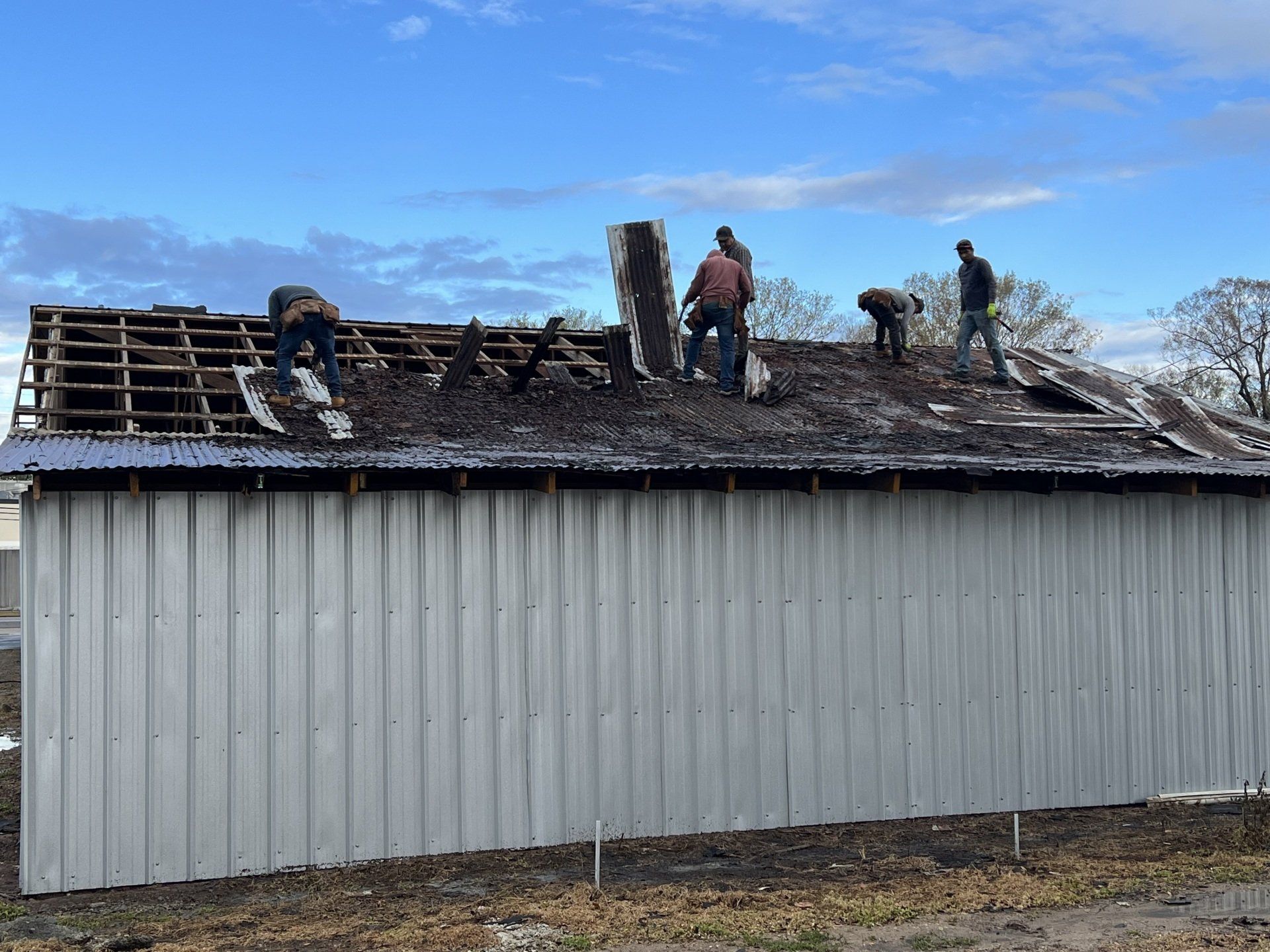 Roofers working on a damaged shingle roof above a white metal-sided building.