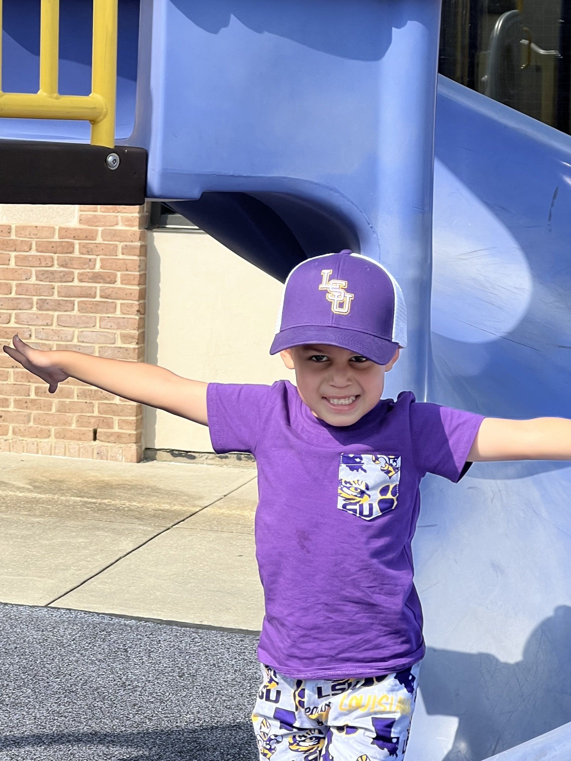 Child in purple shirt and cap, arms outstretched, standing by a blue playground slide.