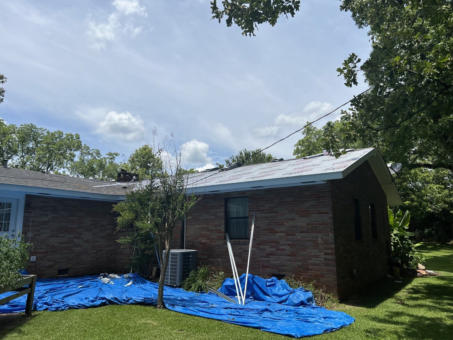 Small brick house with blue tarp spread around the yard and ladders leaning against the wall under a cloudy sky