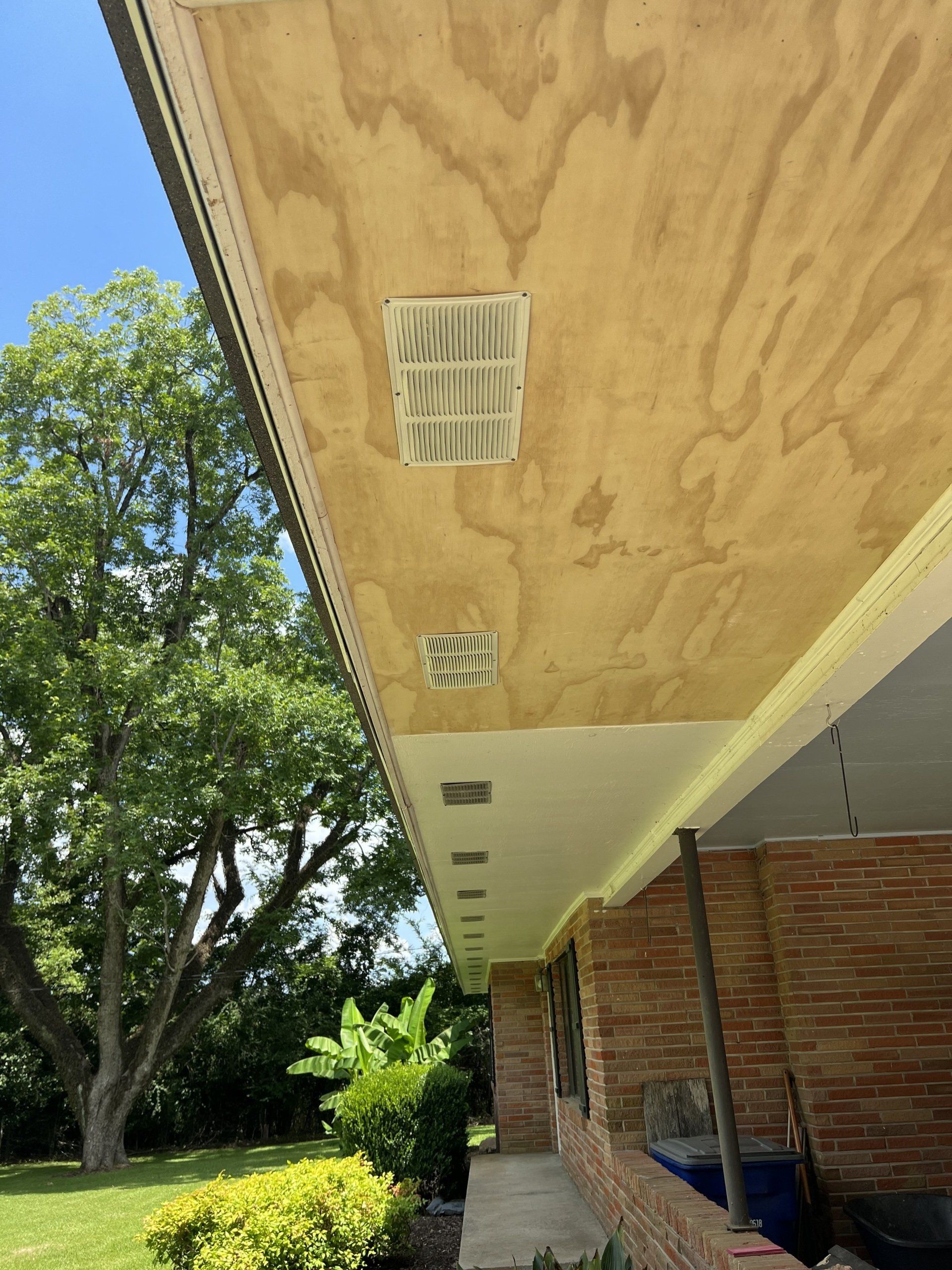 Under a wooden porch roof with vents, beside a brick house and green trees outdoors