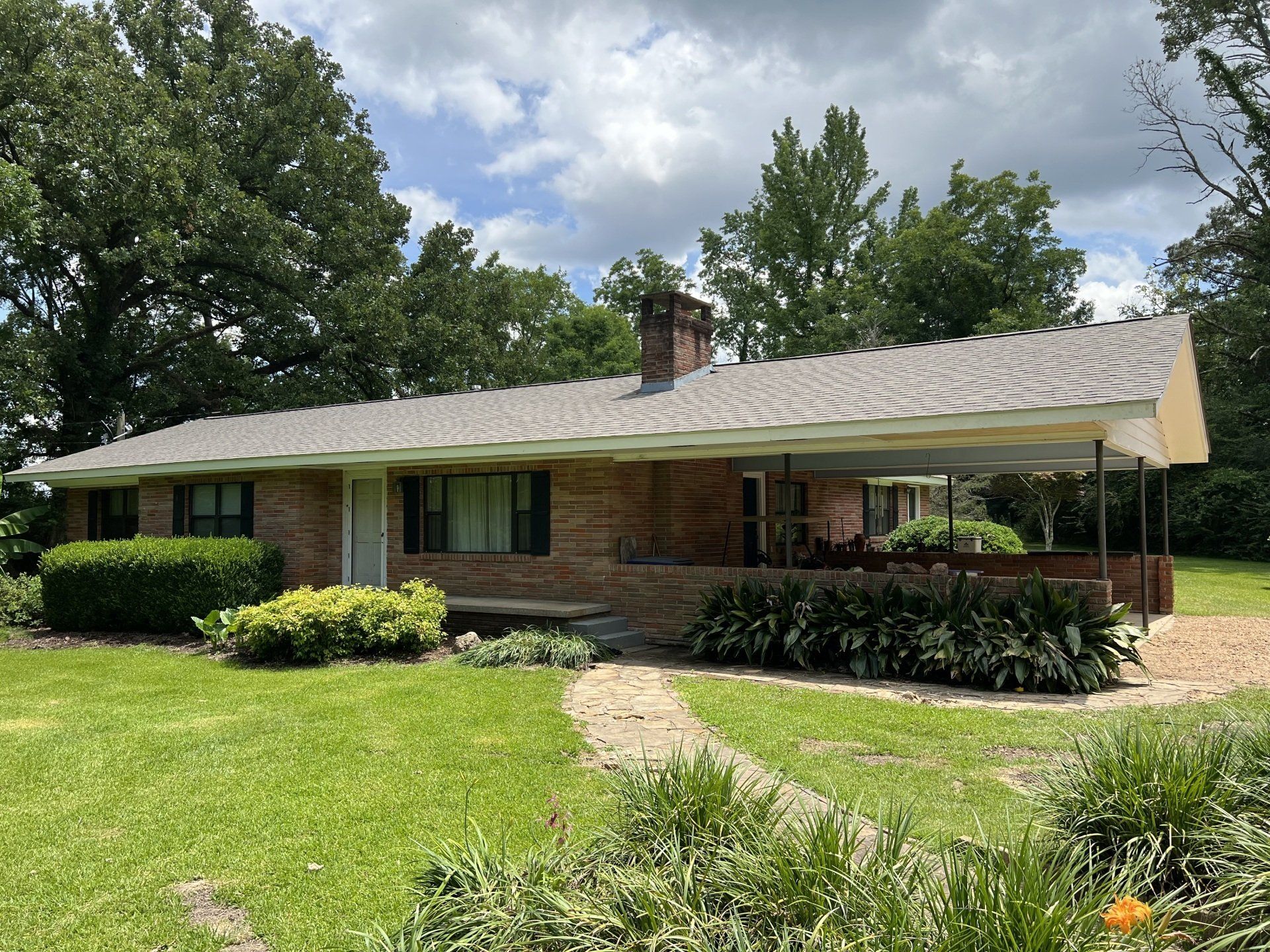 Single-story brick house with a white carport, green lawn, and trees under a cloudy sky
