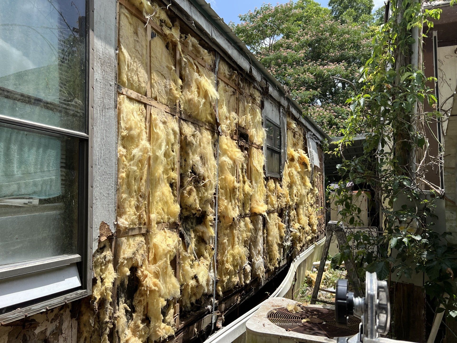 Weathered house wall with exposed yellow insulation beside a window and trees outside