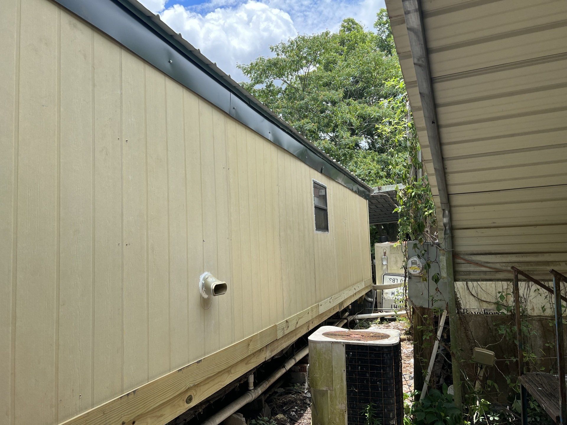 Narrow outdoor passage between a beige mobile home and a covered structure, with trees at the end.