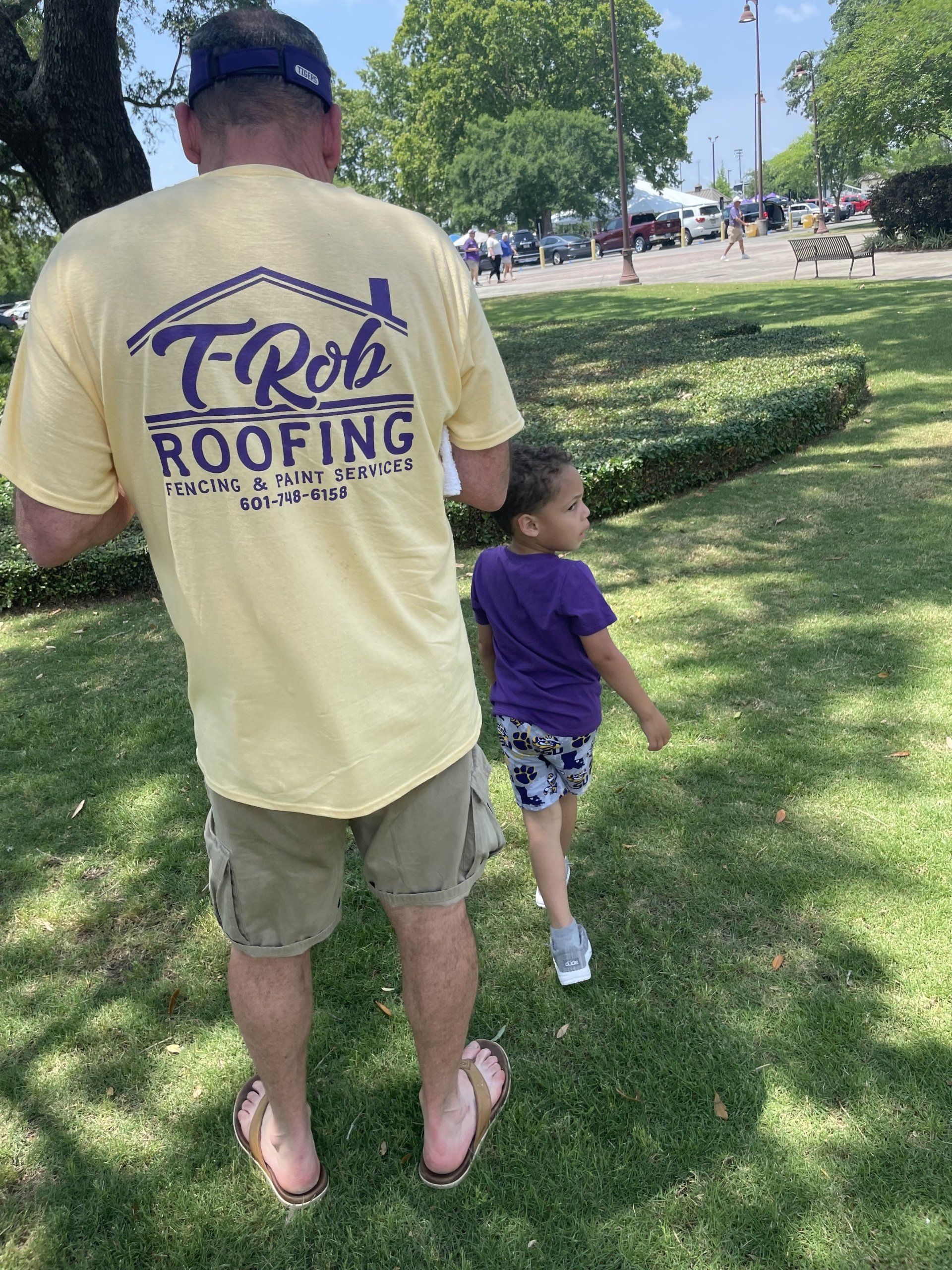 Man in yellow roofing shirt walks beside a child on a sunny park path