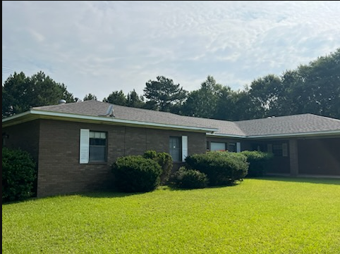 Brick house with green lawn, shrubs, and an attached carport under a cloudy sky