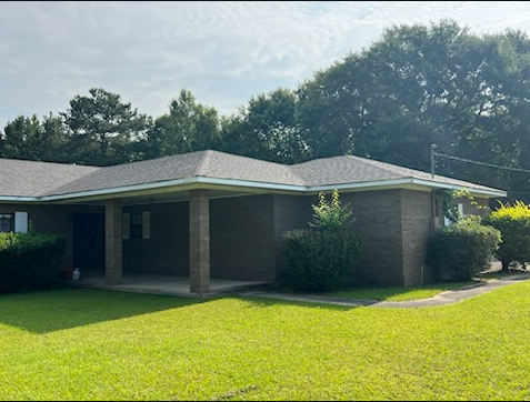 Single-story brick house with a gray roof and green lawn, shaded by trees