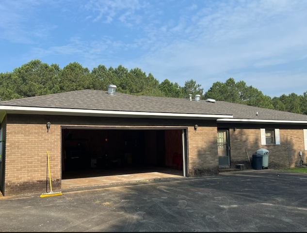 Brick garage building with an open bay door, asphalt driveway, and trees under a blue sky