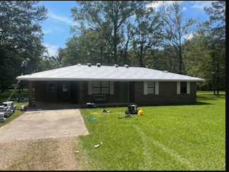 Single-story brick house with a white roof, driveway, and green lawn under trees on a sunny day